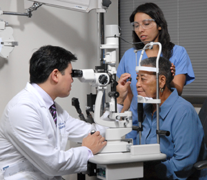 Eye care provider examining person's eye with lens and slit lamp while another provider holds patient's head steady.